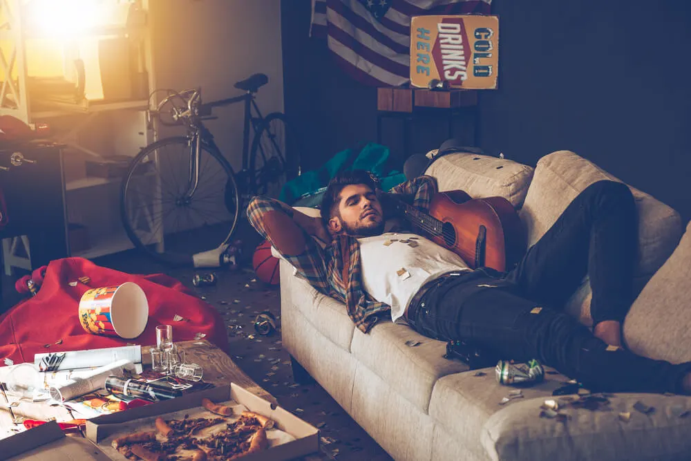 Young handsome man lying down on sofa and napping in messy room after party.