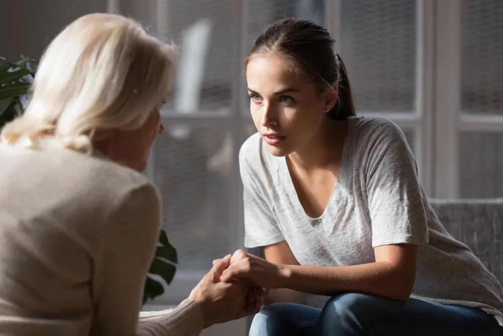 Grown up daughter holds hands of mid aged mother sit on armchair looking at each other having heart-to-heart talk