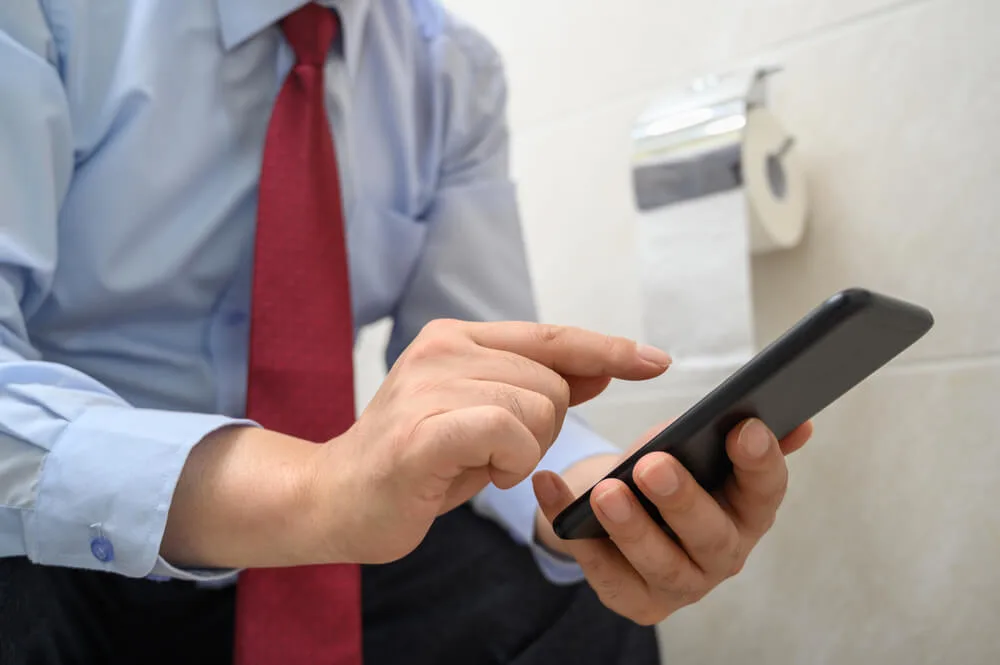 Businessman sitting on the toilet in the bathroom and using a smartphone