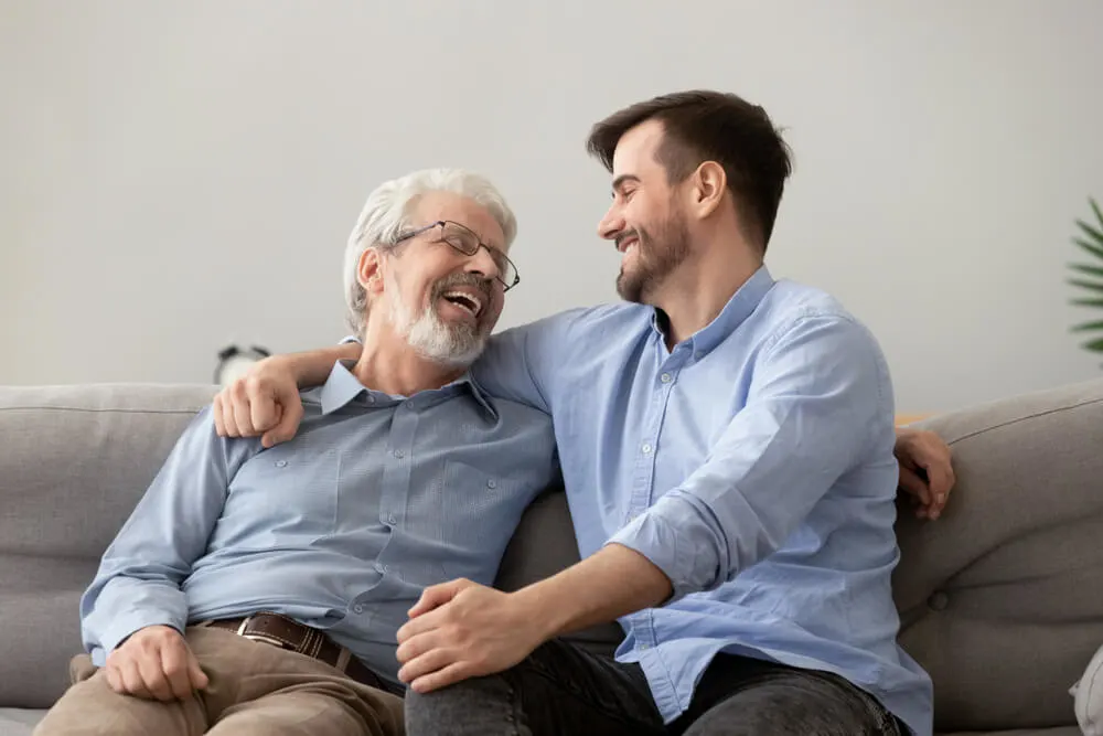 Smiling grown son sit on couch relax with senior dad