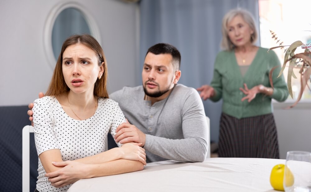 Offended man and his wife sitting at table at home, his mother-in-law standing behind